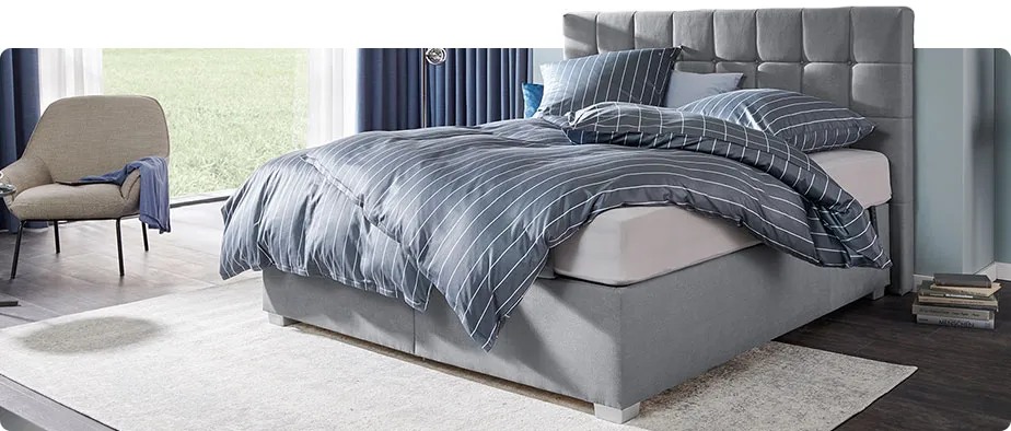 Schlafzimmer mit grauem Boxspringbett mit Matratze und gestepptem Kopfteil - Streifenbettwäsche in Grau, hellgrauer Teppich und beiger Sessel