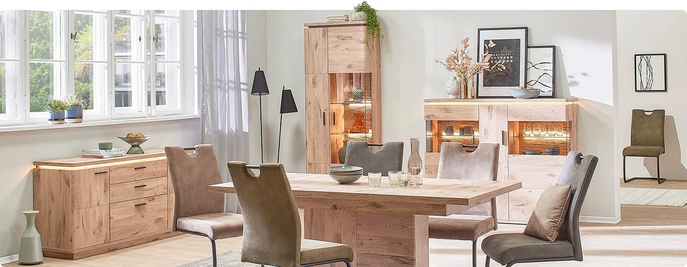 Esszimmer mit Möbeln in Holzoptik, bestehend aus Sideboard, Highboard und Vitrine mit Glas, alle drei mit Beleuchtung sowie Esstisch mit grünen und braunen Polsterstühlen, Deko
