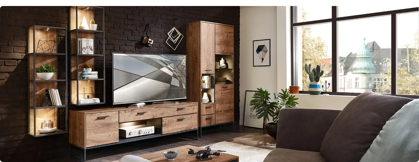 Industrial-Wohnzimmer mit beleuchteter Wohnkombination in Holzoptik mit dunkelgrauen Akzenten bestehend aus Vitrine, Sideboard, 2 Hängeregalen, Wandboard vor dunkelgrauer Steinwand. Im Vordergrund braunes Sofa mit Couchtisch in Holzoptik