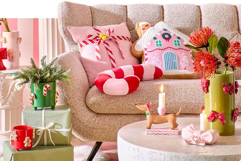 beiges Sofa mit rosa Kissen mit Schleife, rosa-braunes-Kissen in Lebkuchenhaus-Optik, Kissen in rosa-roter Zuckerstangen-Optik, Steintisch mit Dackel-Kerzenhalter, rosa Schleifen-Kerzenhalter, grüner Vase mit roten Schleifen und roten Blumen, Deko-Geschenke in Grün mit rotem und grünem Übertopf