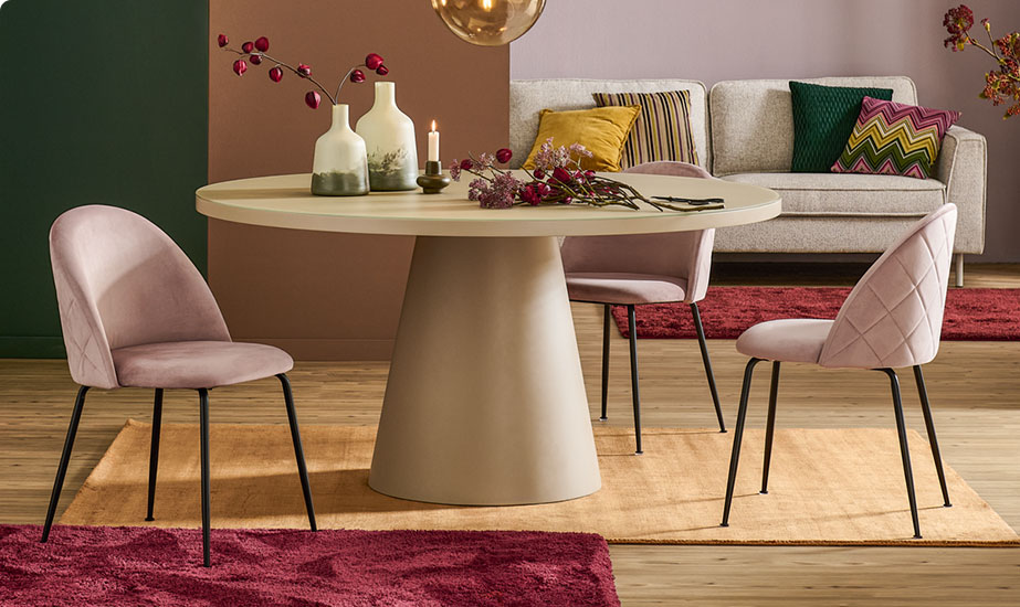 Esszimmer mit beigen, runden Couchtisch mit Vasen und herbstliche Blumen, zwei rosa Polsterstühle, graues Sofa mit bunten Kissen im Hintergrund