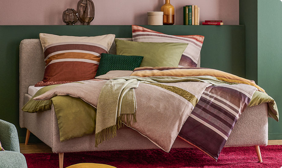Schlafzimmer mit grauem Polsterbett, gestreifte Bettwäsche, Dekokissen und Decken in Grün.