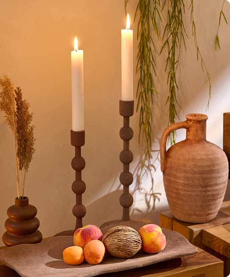zwei braune Kerzenständer mit brennenden Kerzen, braune Steinzeugvase, dunkelbraune kleine Steingutvase, brauner Dekoteller mit Früchten