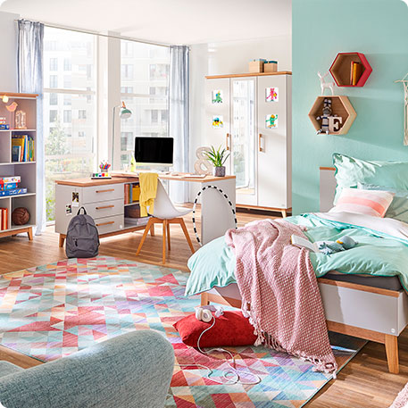 Kinderzimmer mit Bett, Regal, Eckschreibtisch und Schrank in Weiß mit Holzakzenten - bunter Teppich, Stuhl mit gelber Jacke, Rucksack, mintfarbene Bettwäsche und rosa Decke, weitere Deko - im Hintergrund große Fenster