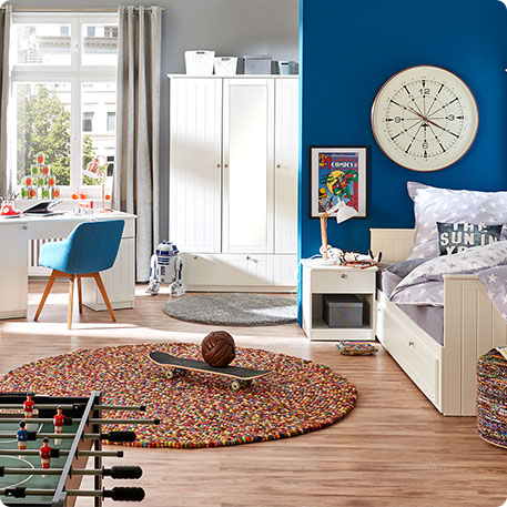 Kinderzimmer mit weißen Möbeln im Landhausstil - Schreibtisch mit blauem Stuhl davor, Schrank und Bett mit grau-weißer Bettwäsche mit Sternen-Muster und grauem Kissen mit weißem Druck "The Sun in You" - Weißer Nachttisch im Landhausstil mit Nachttischlampe vor blauer Wand - Kicker, Teppich und weitere Deko