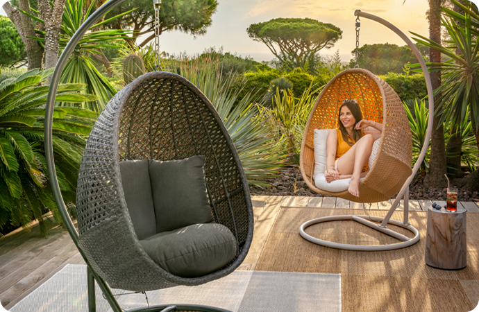 dunkelgrauer Polyrattan-Hängesessel mit dunkler Auflage sowie ein beiger Polyrattan-Hängesessel bei heller Auflage und Frau darin sitzend auf Terrasse vor Palmen 