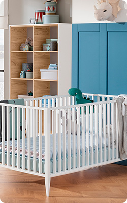 Weißes Babybett mit blauweißem Bettzeug – im Hintergrund ein holzfarbig-weißes Regal und eine blaue Wand