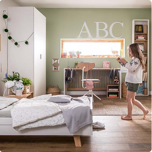Kinderzimmer mit Kinderbett und Schrank. Kind geht durch das Zimmer und hält ein Buch in beiden Händen.