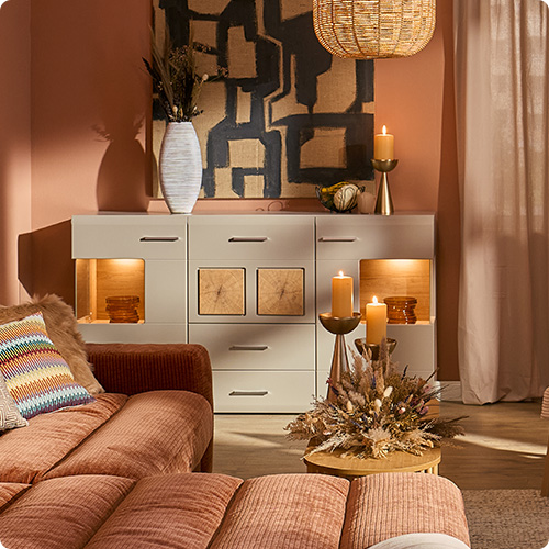 Wohnzimmer mit terracottafarbigem Cord-Ecksofa, weißem Cordsessel mit Holzgestell, Kommode und Vitrine in Grau mit Akzenten in Hirnholzoptik, Deko