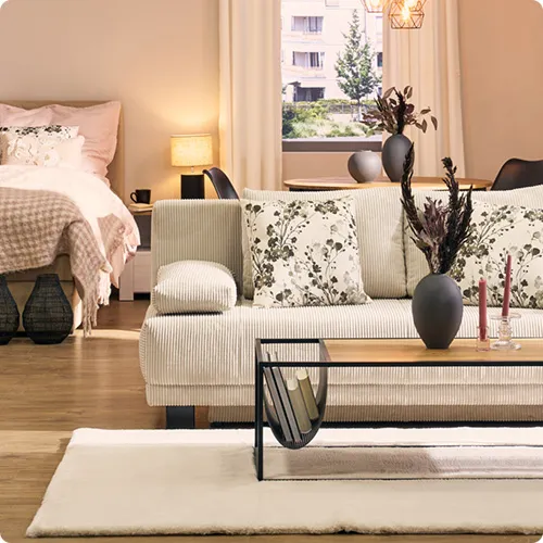 Beiger 2-Sitzer mit geblümten Dekokissen, Holz-Couchtisch mit schwarzem Gestell, weißer Teppich, beiges Bett mit rosa Bettwäsche und Plaid, Deko.