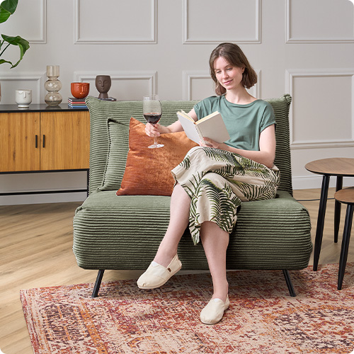 Frau mit Buch sitzt auf grünem Cordsessel, Beistelltische, Teppich im Vintage-Look, Kommode in Holzoptik und Schwarz
