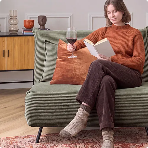Frau mit Buch sitzt auf grünem Cordsessel, Beistelltische, Teppich im Vintage-Look, Kommode in Holzoptik und Schwarz