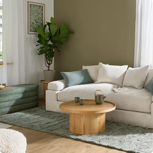 Wohnzimmer mit beigen 2-Sitz-Sofa und 2 grünen Dekokissen. Daneben eine grüne Polsterbank mit Stoffbezug. Vor dem Sofa ein runder Holz-Couchtisch 