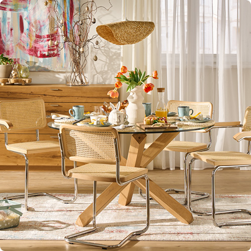 Esszimmer mit gedecktem Esstisch aus Glas und Holz, Schwingstühle aus Ratten, Holz und Metall, Kommode aus Holz im Hintergrund