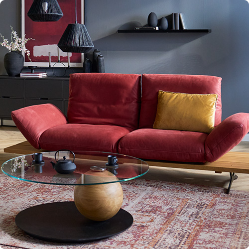 Wohnzimmer mit rotem Ledersofa, davor ein runder Glastisch mit Holz und schwarzem Metall, im Hintergrund eine schwarze Kommode und Wandregal