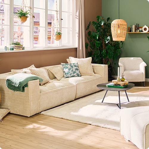 Cremefarbiges Cordsofa, weißer Sessel und Hocker, weißer Teppich, schwarzer Couchtisch, Pflanzen und Deko in einem Wohnzimmer