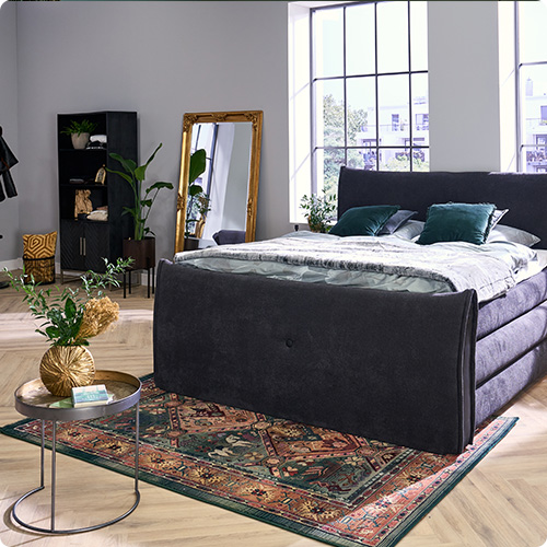 Schwarzes Boxspringbett mit grauer Bettwäsche und grünen Kissen, schwarze Möbel und goldener Spiegel in einem Schlafzimmer