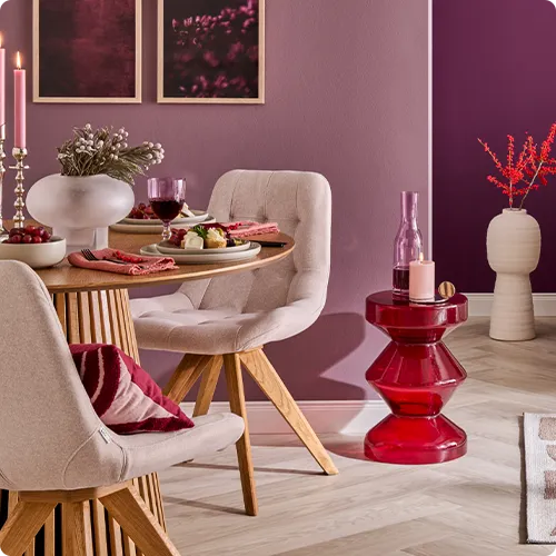 Gedeckter runder Holztisch mit 2 cremefarbigen Polsterstühlen, roter Glasttisch mit Deko, cremefarbige Bodenvase, lila Wände mit Bildern