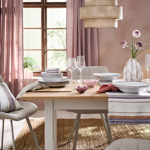Farben der Provence - Sommertrend 2026. Gedeckter Holztisch mit 2 grauen Polsterstühlen, gestreifter Tischläufer, Platzsets, graues Geschirr mit dunklem Rand, Weingläser, Blumenvase und Teelichthaltern in Blütenform, Pendelleuchte mit Stoffschirm