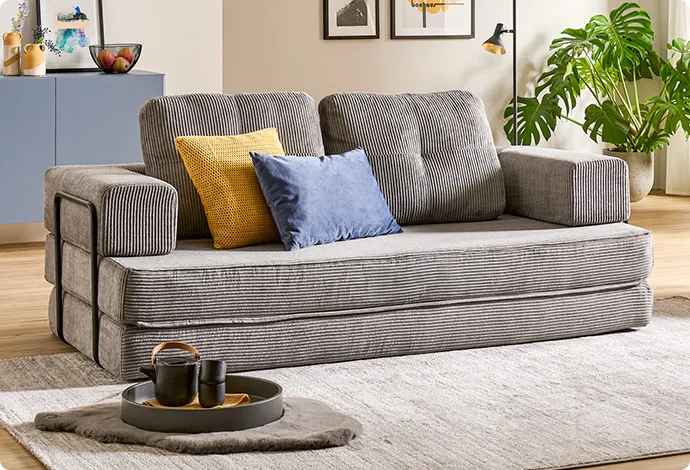 Wohnzimmer mit grauem Cordsofa, gelbem und blauem Kissen, grauem, Sideboard, Teppichen und Dekoration