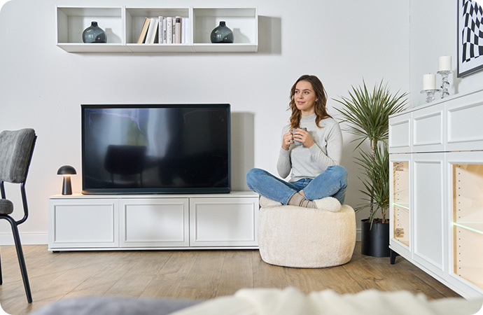 TV-Lowboard und Sideboard, beide weiß und im Landhausstil - Sideboard mit 2 Vitrinenfächern, beleuchtet - Frau mit Kaffeetasse, die auf cremefarbigem Pouf sitzt