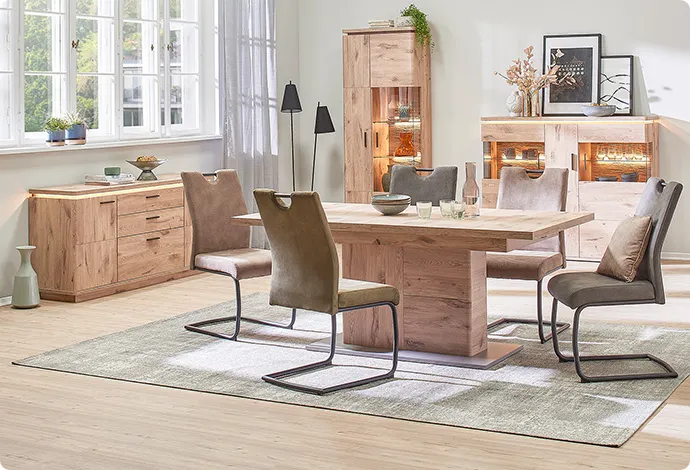 Esszimmer mit Möbeln in Holzoptik, bestehend aus Sideboard, Highboard und Vitrine mit Glas, alle drei mit Beleuchtung sowie Esstisch mit grünen und braunen Polsterstühlen, Deko