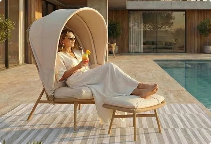 Frau sitzt auf einem Lounge-Sessel mit Sonnendach und hat die Füße auf einem Hocker liegen. Sie hält ein Getränk in der Hand.