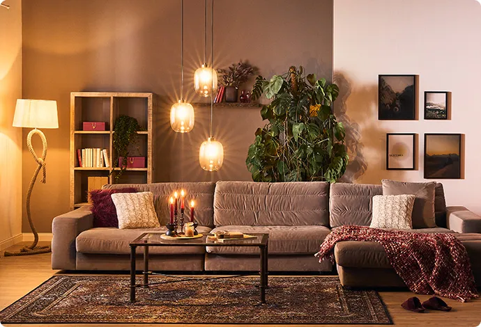 Wohnzimmer mit großem braunen Sofa, Kerzen auf dem Couchtisch, einer Steh- und einer Pendelleuchte. Bilder hängen an der Wand. Eine große Pflanze steht im Hintergrund.