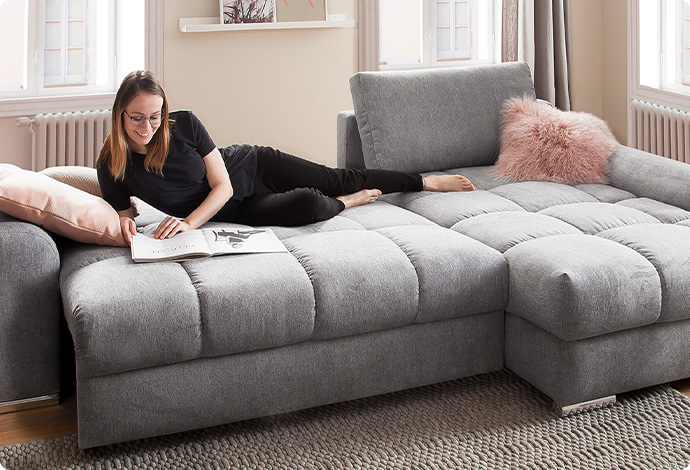 Graues Schlafsofa in L-Form mit darauf liegender, lesender Frau in einem Wohnzimmer
