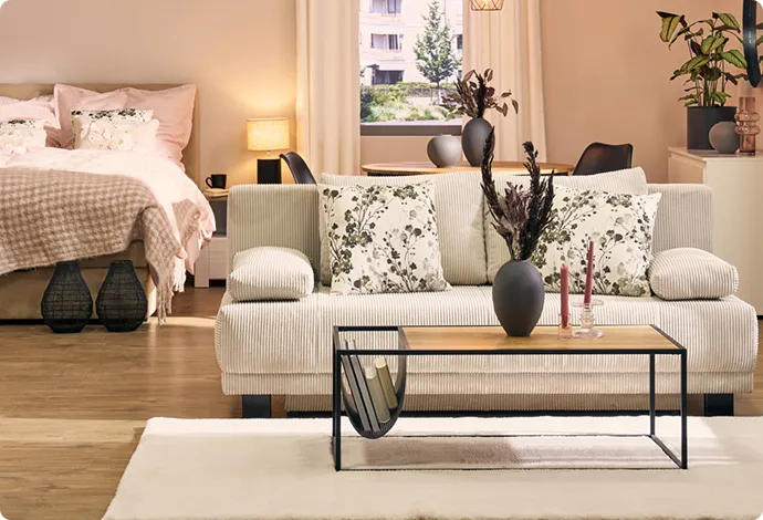 Beiger 2-Sitzer mit geblümten Dekokissen, Holz-Couchtisch mit schwarzem Gestell, weißer Teppich, beiges Bett mit rosa Bettwäsche und Plaid, Deko.
