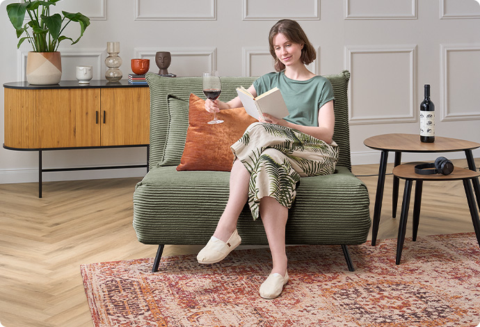 Frau mit Buch sitzt auf grünem Cordsessel, Beistelltische, Teppich im Vintage-Look, Kommode in Holzoptik und Schwarz