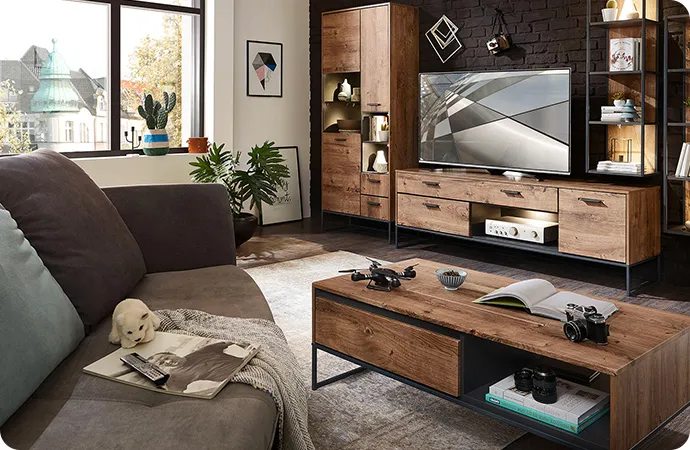 Industrial-Wohnzimmer mit beleuchteter Wohnkombination in Holzoptik mit dunkelgrauen Akzenten bestehend aus Vitrine, Sideboard, 2 Hängeregalen, Wandboard vor dunkelgrauer Steinwand. Im Vordergrund braunes Sofa mit Couchtisch in Holzoptik