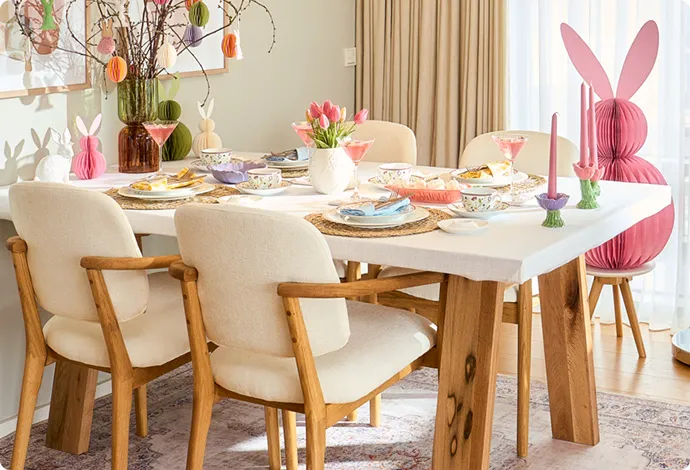 Esstisch mit Polsterstühle in Weiß mit Holzfüßen, große und kleine Deko-Papierosterhasen in Wabenoptik, Äste mit Eier-und Osterhastenanhänger aus Papier in Pastelltönen, geflochtene runde Platzsets, blumiges Geschirr, Kerzenständer in Tulpenform und Pastellfarben, Dekoschalen in Pastell. 