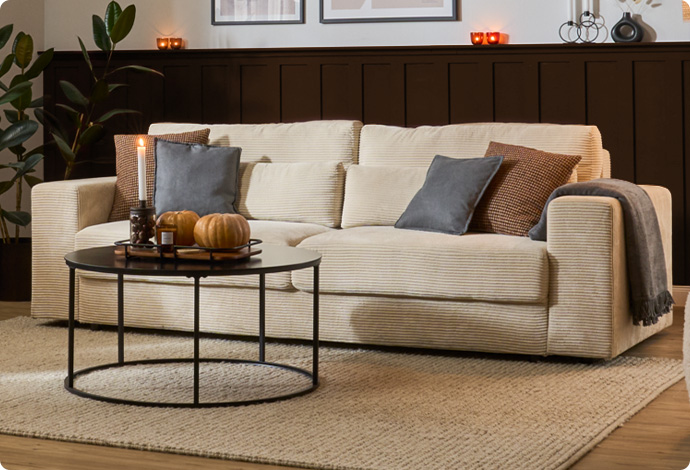 Wohnsituation: cremefarbiges Cordsofa mit diversen Kissen und Decke, schwarzer Couchtisch mit Deko in einem Wohnzimmer.