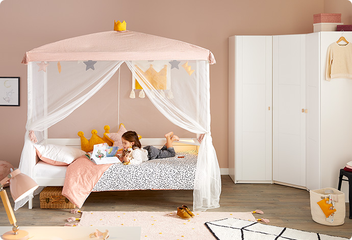 Weißes Kinderbett mit Baldachin in Weiß und Rosa mit gelbem Krönchen, lesendes Mädchen in Bett, Bettwäsche, Kissen, Spielzeug und Deko