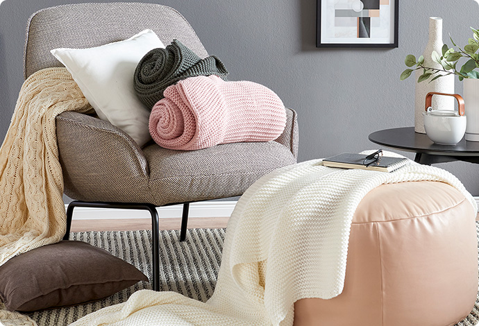 Beiger-brauner Sessel mit weißem Kissen und 2 Strickdecken in Rosa und Grün sowie rosa Pouf mit Strickdecke in Weiß. Eine beige Strickdecke liegt über der Sessellehne. Ein braunes Kissen liegt auf dem Boden.