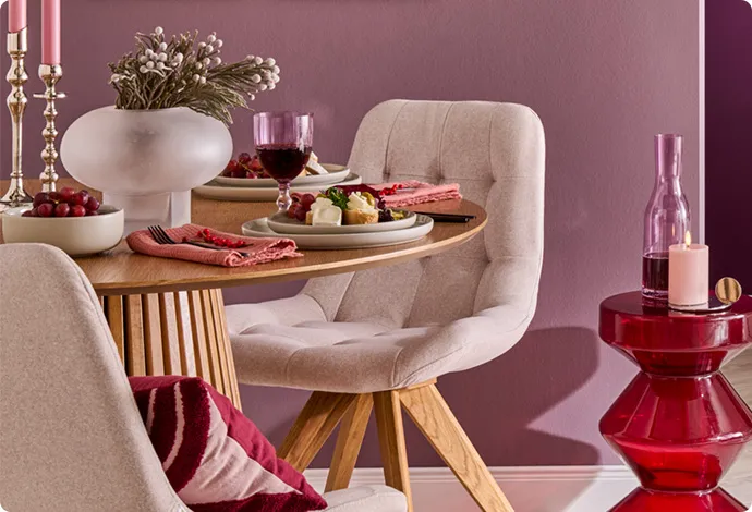 Gedeckter runder Holztisch mit 2 cremefarbigen Polsterstühlen, roter Glasttisch mit Deko, cremefarbige Bodenvase, lila Wände mit Bildern