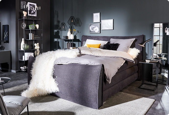 graues Boxspringbett mit hellgrauer Bettwäsche, weißer Felldecke, schwarzem und gelbem Kissen in einem grauen Schlafzimmer mit schwarzen Beimöbeln und Deko