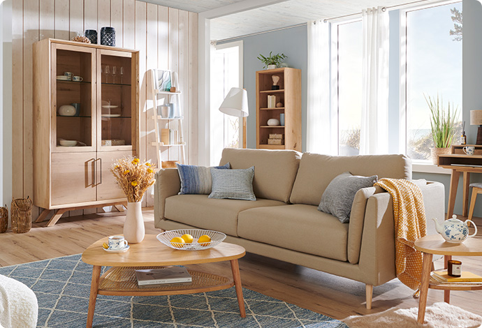 Wohnzimmer mit beigen Sofa, Holz-Couchtisch auf blauem Teppich mit weißem Muster, im Hintergrund Vitrine und Regal aus Holz, Deko