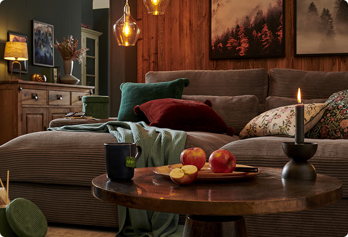 Graues Cordsofa, grünes und rostbraunes Kissen, grüne Decke, brauner runder Couchtisch in einem Wohnzimmer mit Holzwand