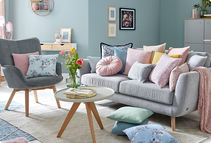 Graues Sofa mit vielen pastellfarbigen Kissen, runder Couchtisch in Grau mit Holzgestell in einem Wohnzimmer