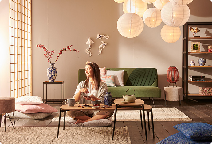 Frau in einem asiatisch eingerichtetem Wohnzimmer mit 2 Couchtischen aus Holz und Metall, grünem Sofa, weißen Pendelleuchten und weiteren Möbeln und Deko