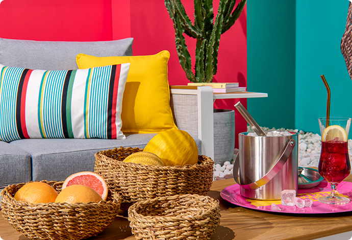 3 braune Körbe, 2 davon mit Obst, auf einem Holztisch, graues Gartensofa mit einem bunt-gestreiften und einem gelben Kissen, Kaktus vor pinker Wand 