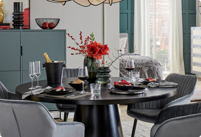 schwarzer runder Tisch, gedeckt, graue Polsterstühle mit schwarzen Beinen, graue Kommode im Hintergrund