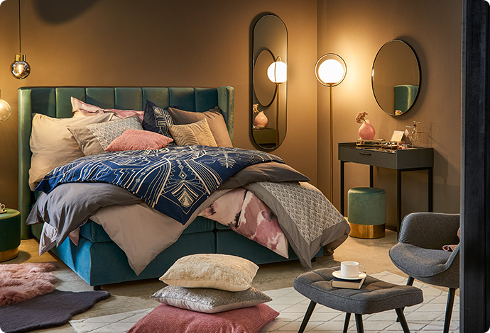 3 aufeinander gestapelte Kissen unterschiedlicher Größe in Rot, Grau und Beige, grauer Hocker, weißer Teppich, Bett mit mehrfarbiger, gemusterter Bettwäsche in Schlafzimmer