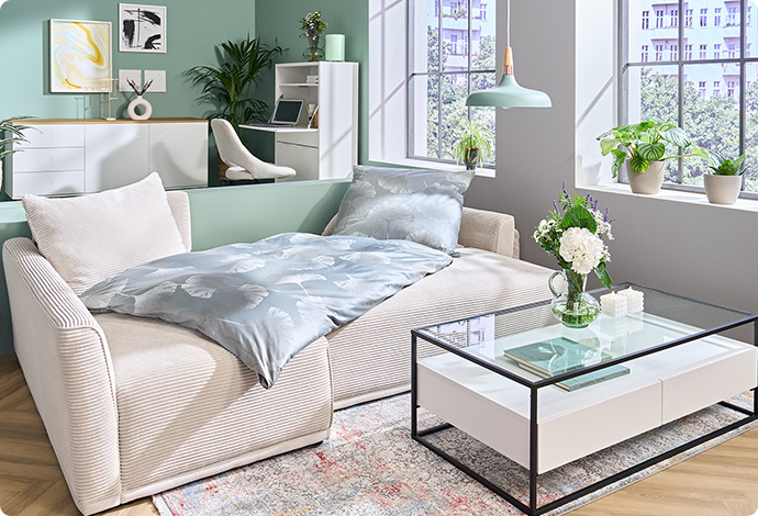 Single-Appartement mit ausgeklappter Schlafcouch aus Cord mit grauem Kopfkissen und grauer Bettdecke, Glas-Couchtisch und weitere Möbel