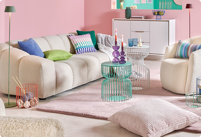 Wohnzimmer mit weißem Sofa, weißem Sessel und weißer Kommode, Metall-Couchtische in Mintgrün und Weiß, Textilien und Deko in Pastelltönen, rosa Wände