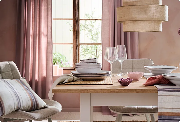 Farben der Provence - Sommertrend 2026. Gedeckter Holztisch mit 2 grauen Polsterstühlen, gestreifter Tischläufer, Platzsets, graues Geschirr mit dunklem Rand, Weingläser, Blumenvase und Teelichthaltern in Blütenform, Pendelleuchte mit Stoffschirm