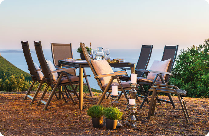 gedeckter Gartentisch, 6 Klappstühle und passender Hocker, alle aus grauem Alu und Textilgewebe in einem Garten vor dem Meer
