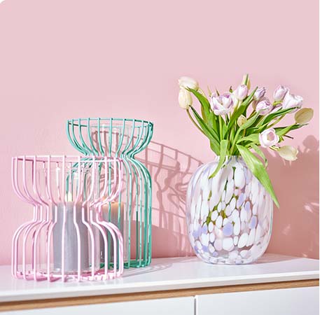 Zwei Windlichter aus Draht in Rosa und Türkis, daneben eine Glasvase mit weißen und Lila Tupfen mit Tulpenstrauß dekoriert. Alles steht vor einer rosa Wand